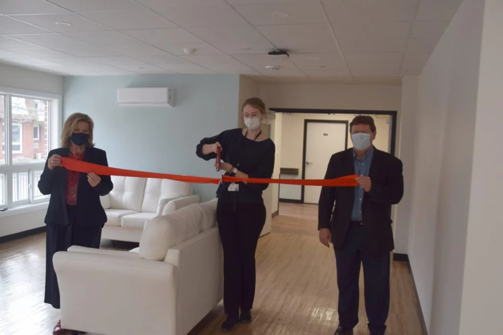 Three people wearing masks cut a ribbon to mark the opening of a long-term care facility.