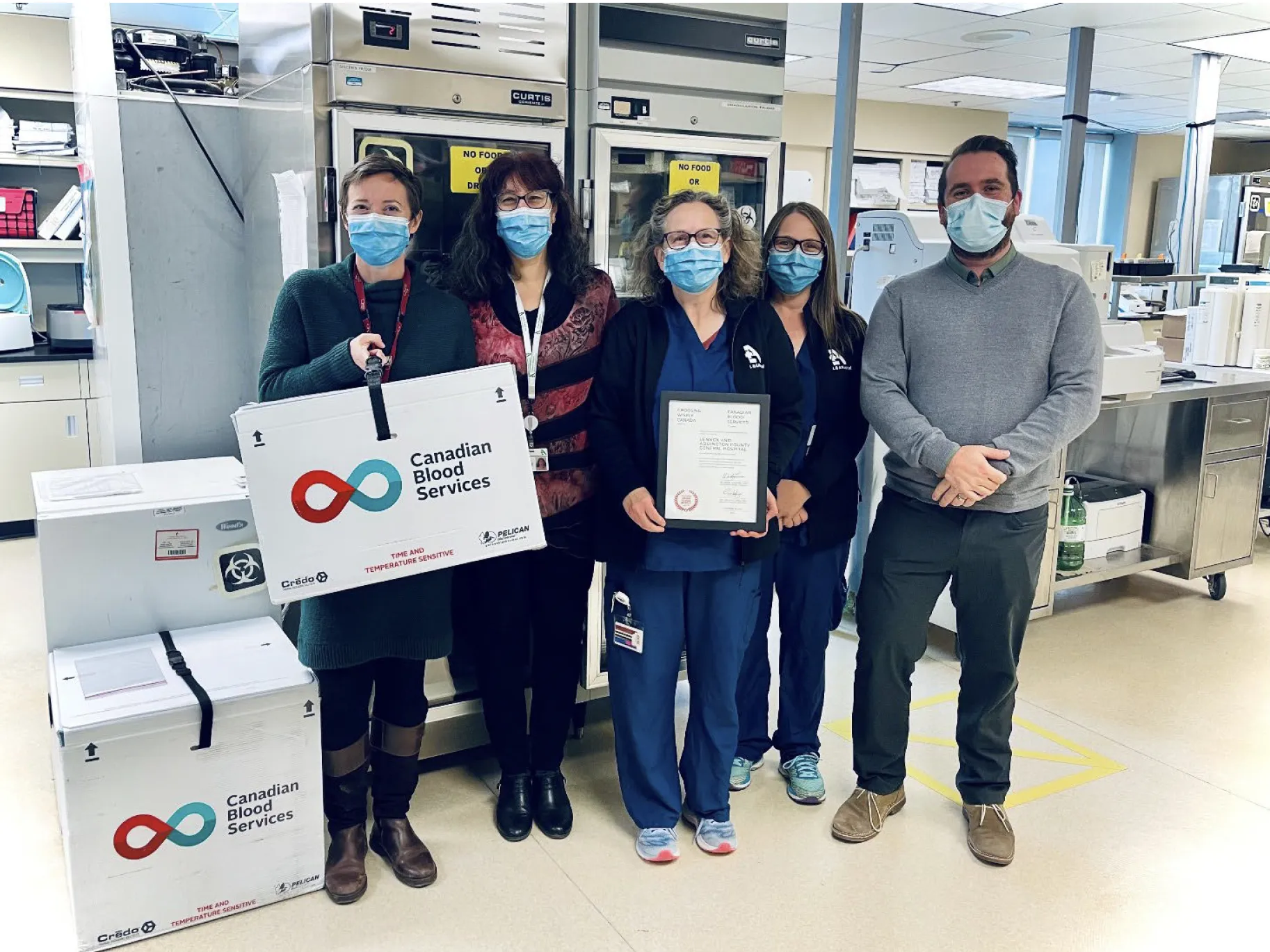 Group of hospital staff wearing masks standing indoors, one person holding a framed certificate beside Canadian Blood Services containers.