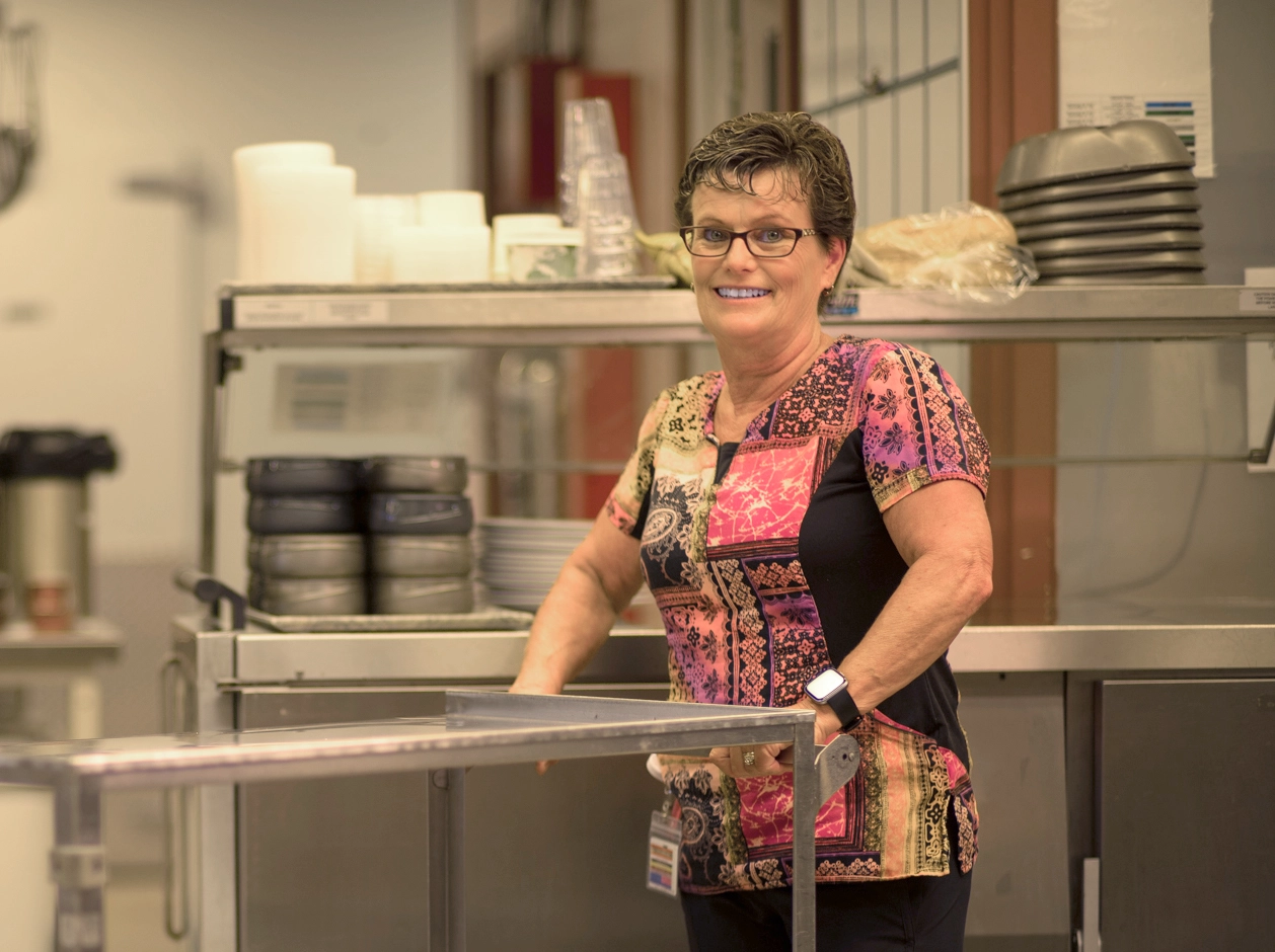 Food service worker stands behind a stainless-steel counter in a hospital kitchen, smiling toward the camera.