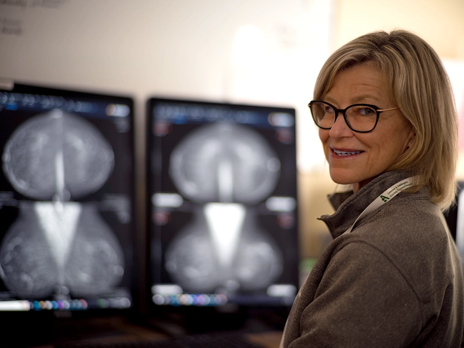 Health care professional smiling in front of dual medical imaging monitors displaying diagnostic scans.