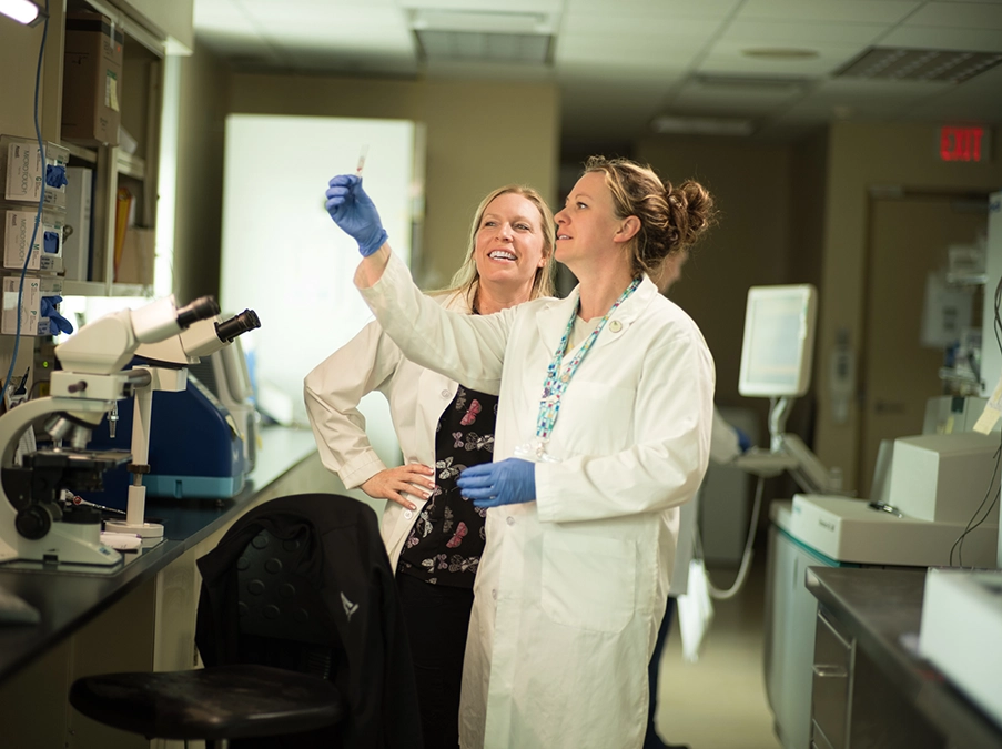 Two laboratory professionals in white coats examining a test sample together in a clinical lab.