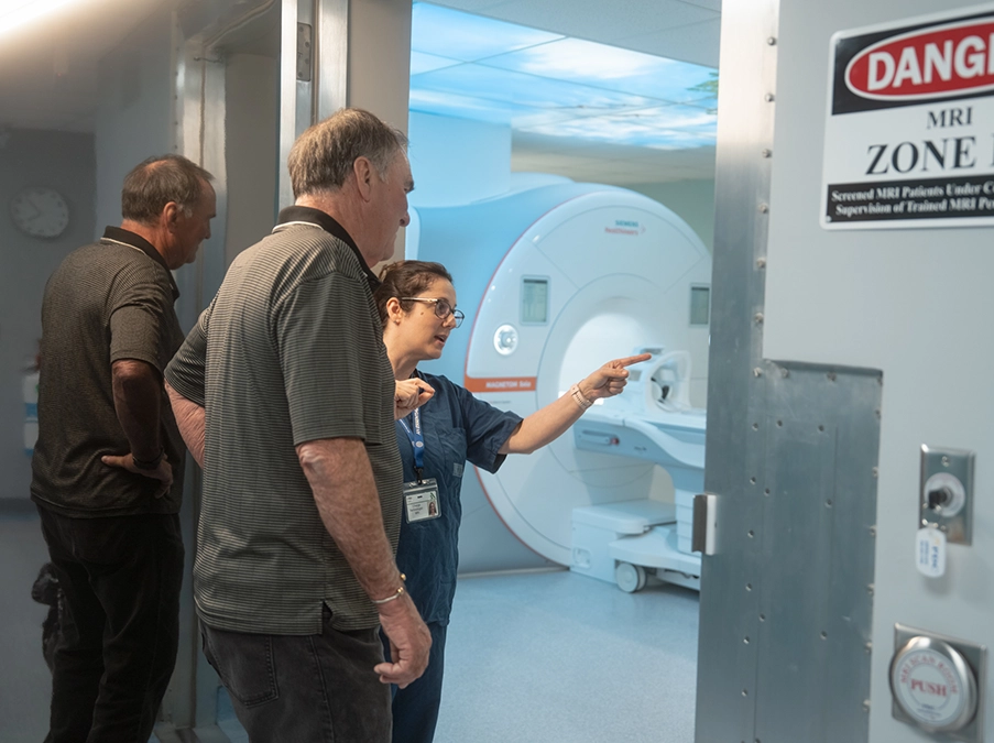 Health-care worker points toward an MRI scanner while explaining the procedure to two patients outside the MRI room.