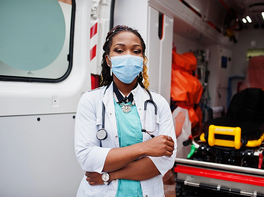 Masked health care worker standing inside an ambulance with medical equipment visible behind them.