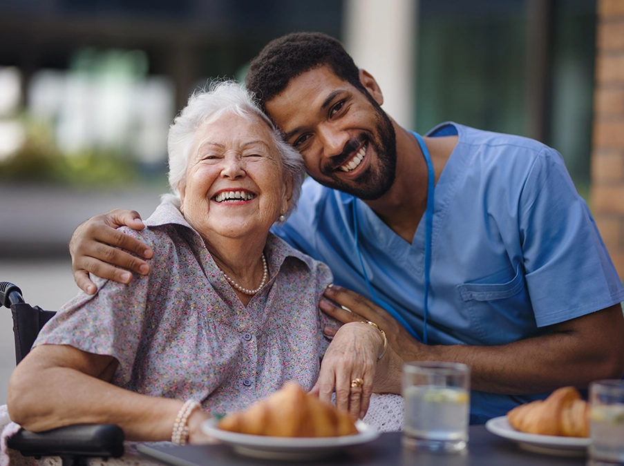 Health care worker and elderly patient smiling together at a table with food, showing a supportive caregiver relationship.