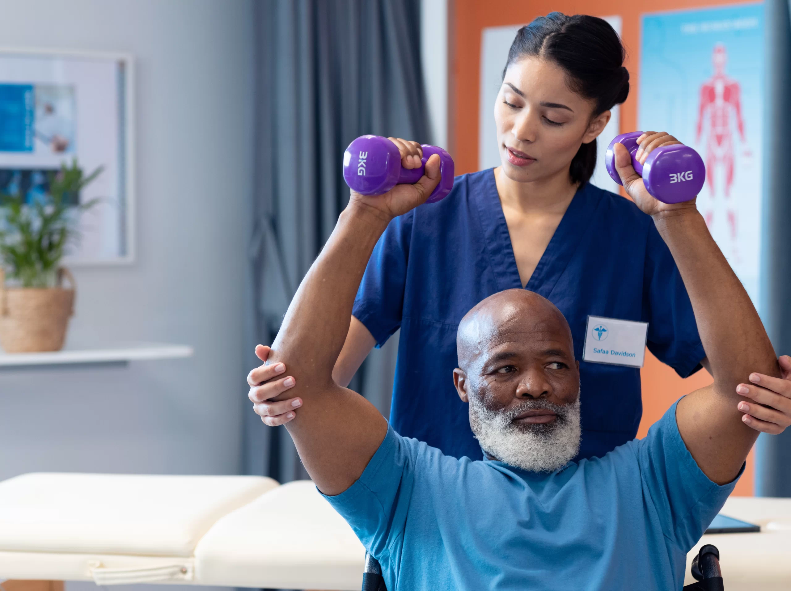 Physiotherapist supporting an older man as he lifts two purple hand weights during a seated exercise session.