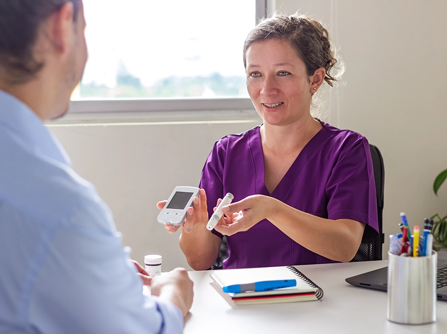 Nurse in purple scrubs demonstrates a blood glucose monitor and test strip to a seated patient during a consultation.