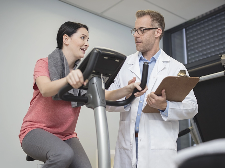 Clinician discussing treatment progress with a patient using a stationary exercise bike in a rehabilitation setting.