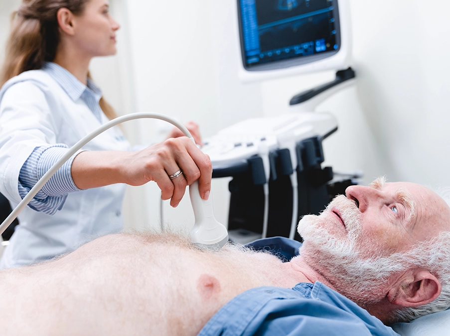 Clinician performs an ultrasound scan on an older patient lying on an exam table while viewing the monitor.