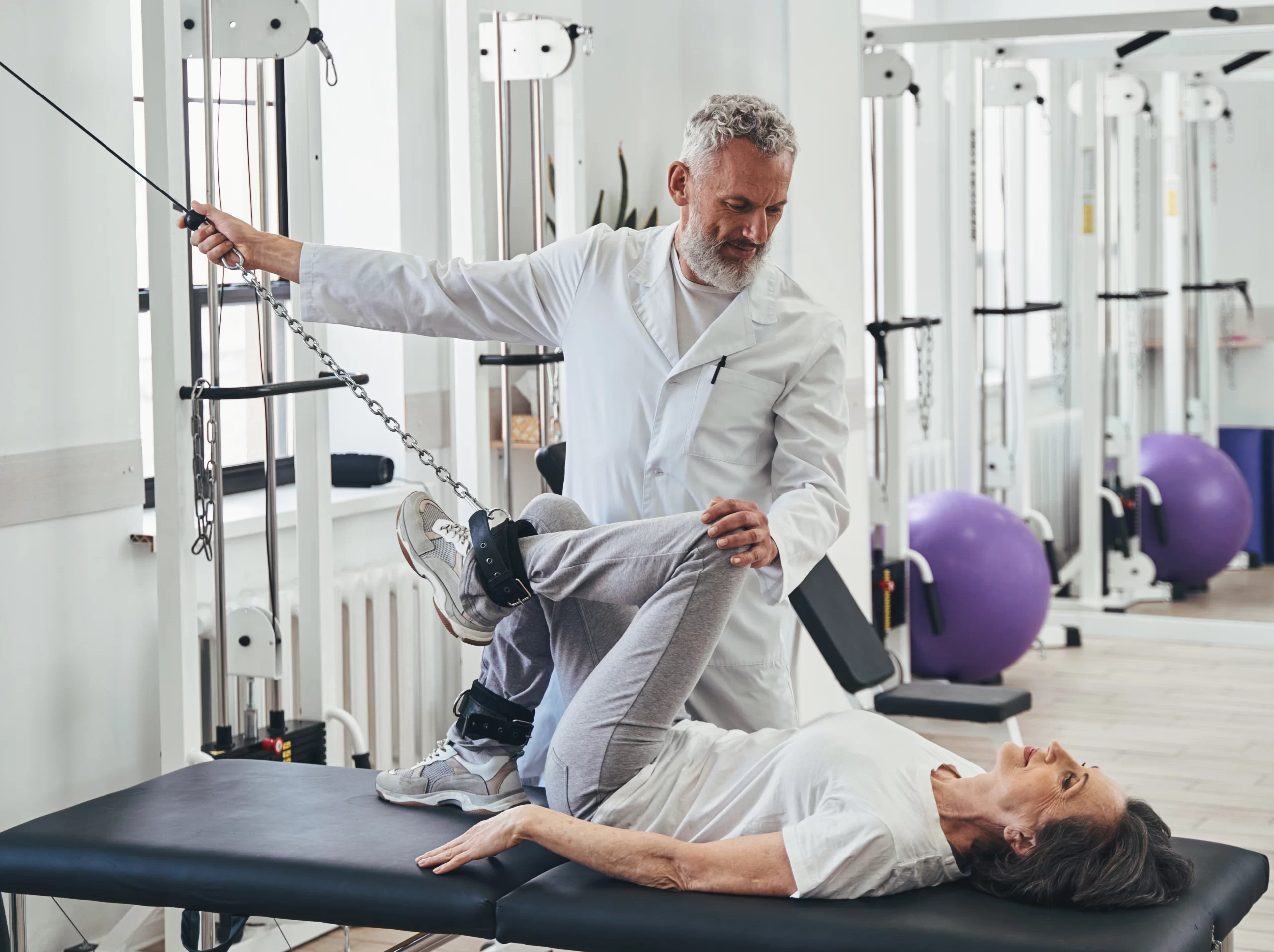Physiotherapist assists an older patient with a leg exercise on a treatment table in a rehabilitation gym.