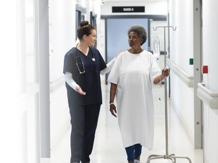 Health-care worker walking alongside an older patient in a hospital hallway, offering support while the patient holds an IV stand.