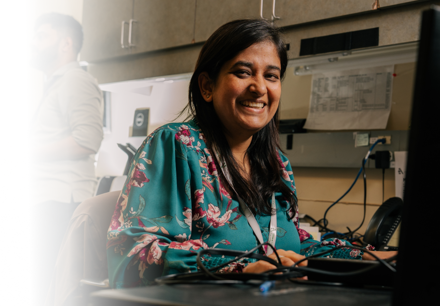 Staff member smiling while working at a computer workstation in an office, with a colleague working in the background.