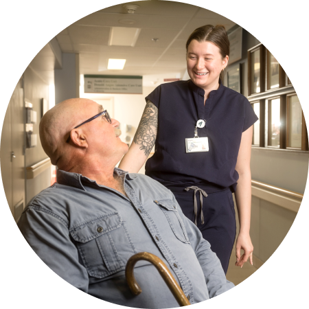 Health-care worker smiling and speaking with an older adult using a walker in a hospital corridor.