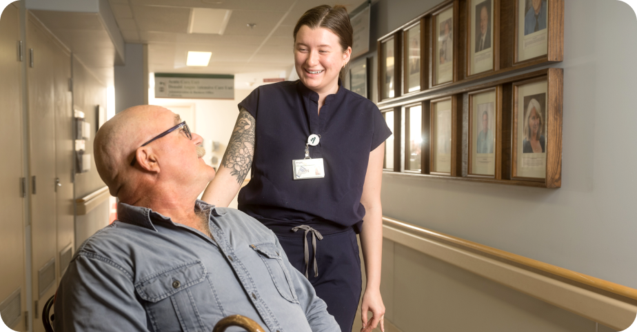 Health-care worker smiling and speaking with an older adult using a walker in a hospital corridor.