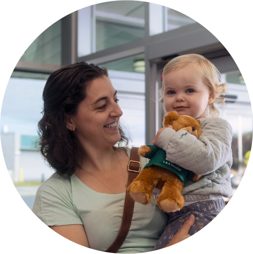 An adult holding a toddler indoors, with the child holding a stuffed toy and looking toward the camera.