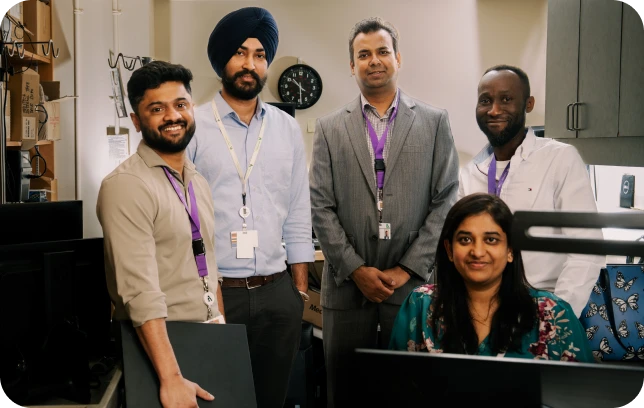 Five staff members standing together in an office workspace, smiling toward the camera, with desks and computer equipment in the background.