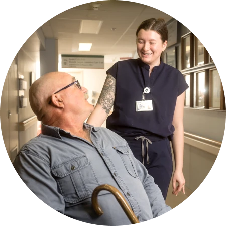 Health-care worker smiling and speaking with an older adult using a walker in a hospital corridor.