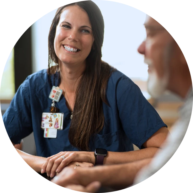 A nurse in scrubs speaking with an older adult at a table in a health-care setting.”