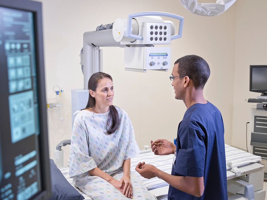 Clinician speaks with a patient seated on an exam table beneath an overhead medical imaging device.