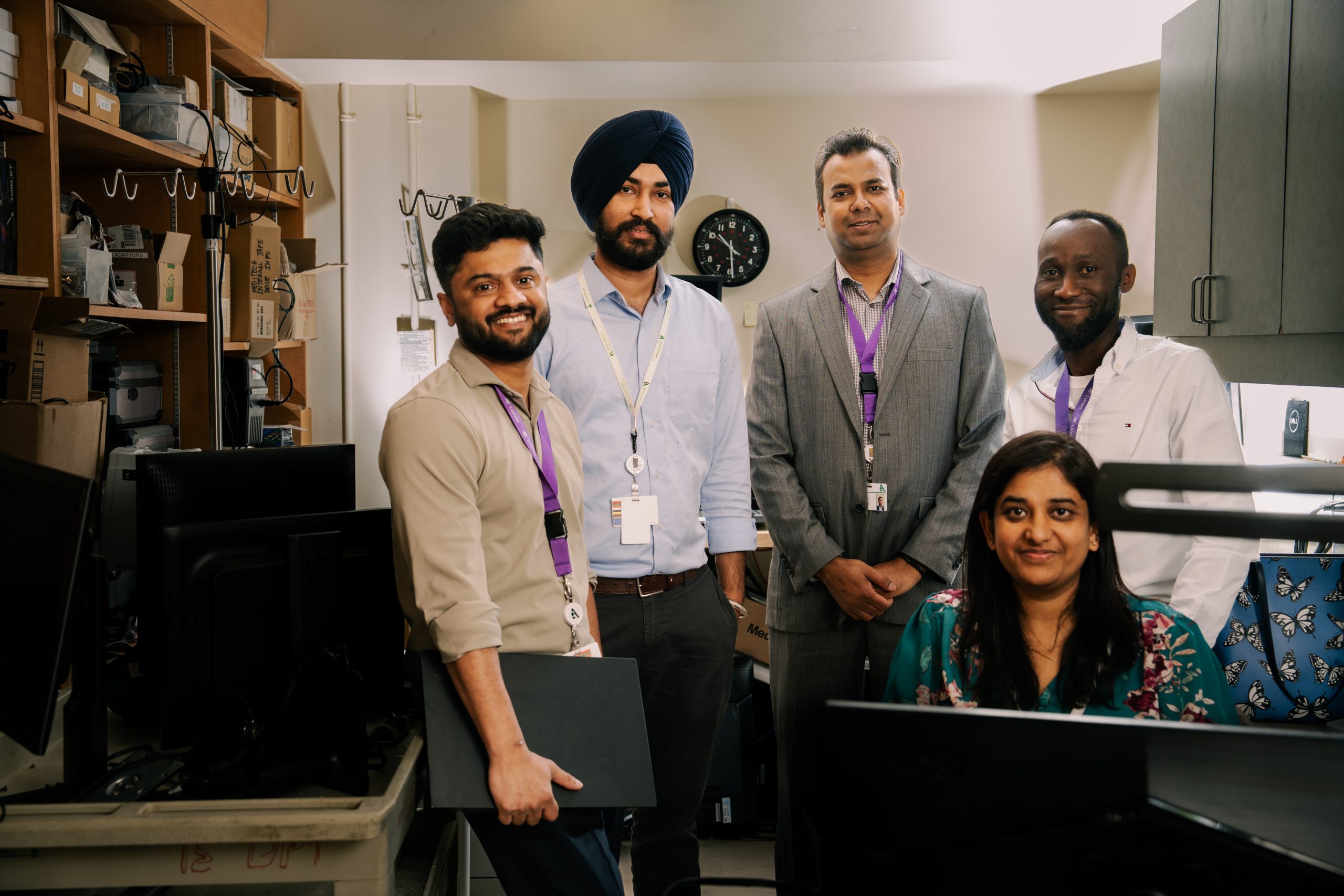 Five staff members standing together in an office workspace, smiling toward the camera, with desks and computer equipment in the background.