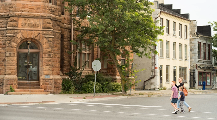 Adult and two children walk across a street in front of older brick and stone buildings, including a former post office.