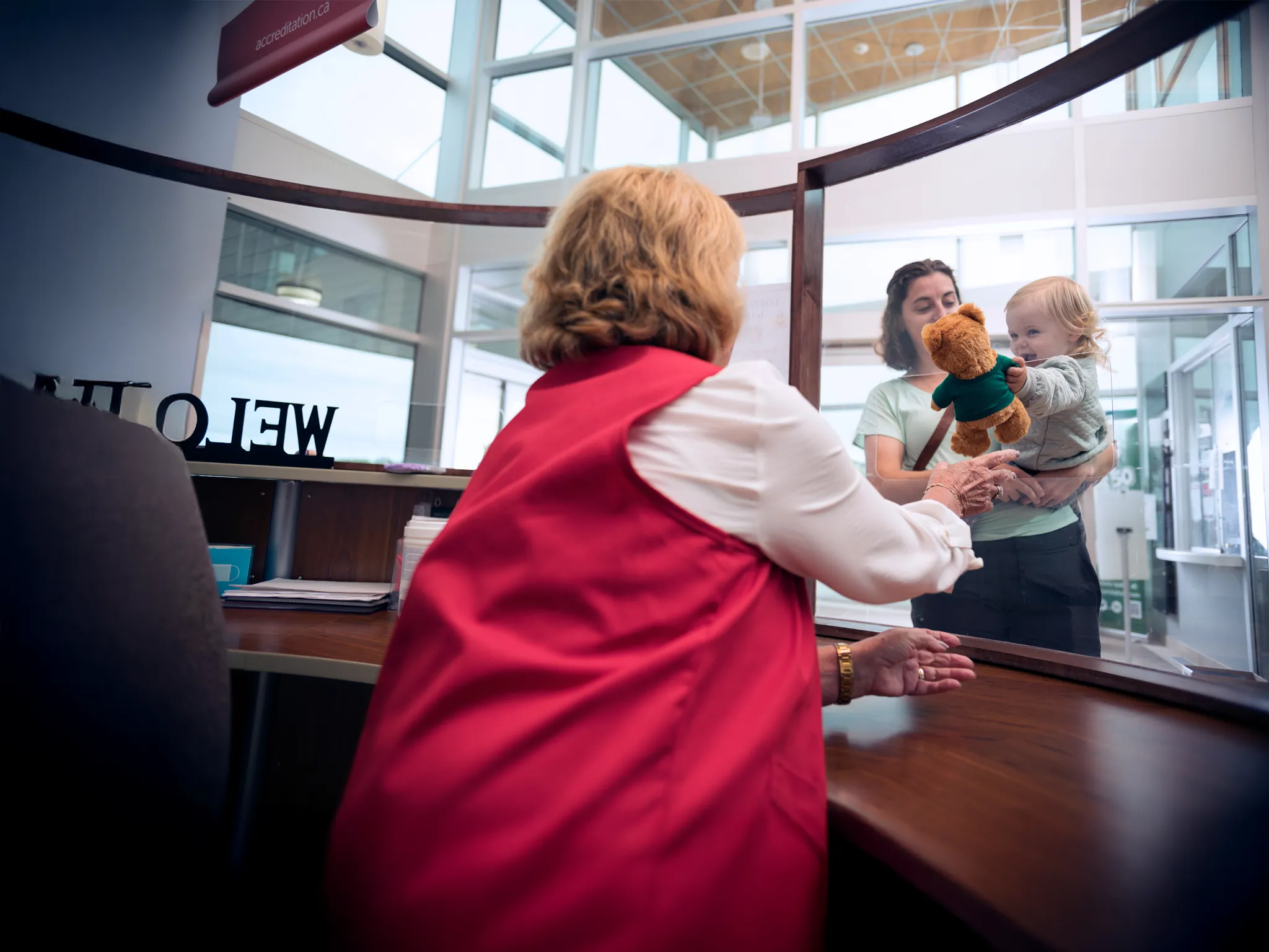 Hospital volunteer at an information desk hands a teddy bear to a child.