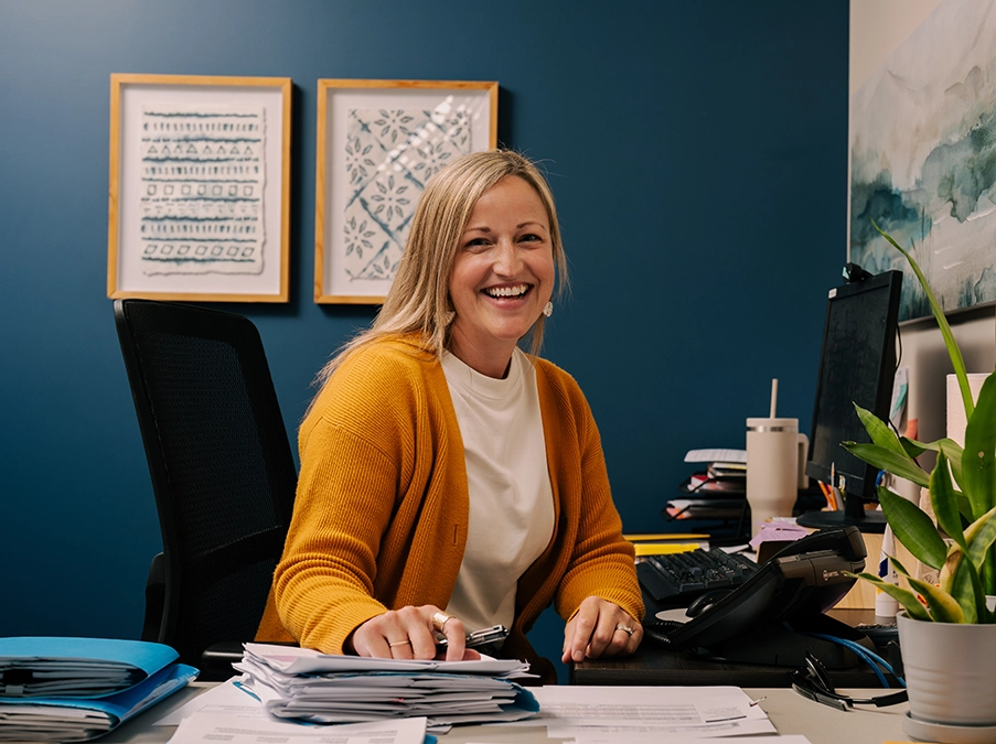 Office staff member seated at a desk, smiling toward the camera, with paperwork, a computer, and a potted plant on the desk.