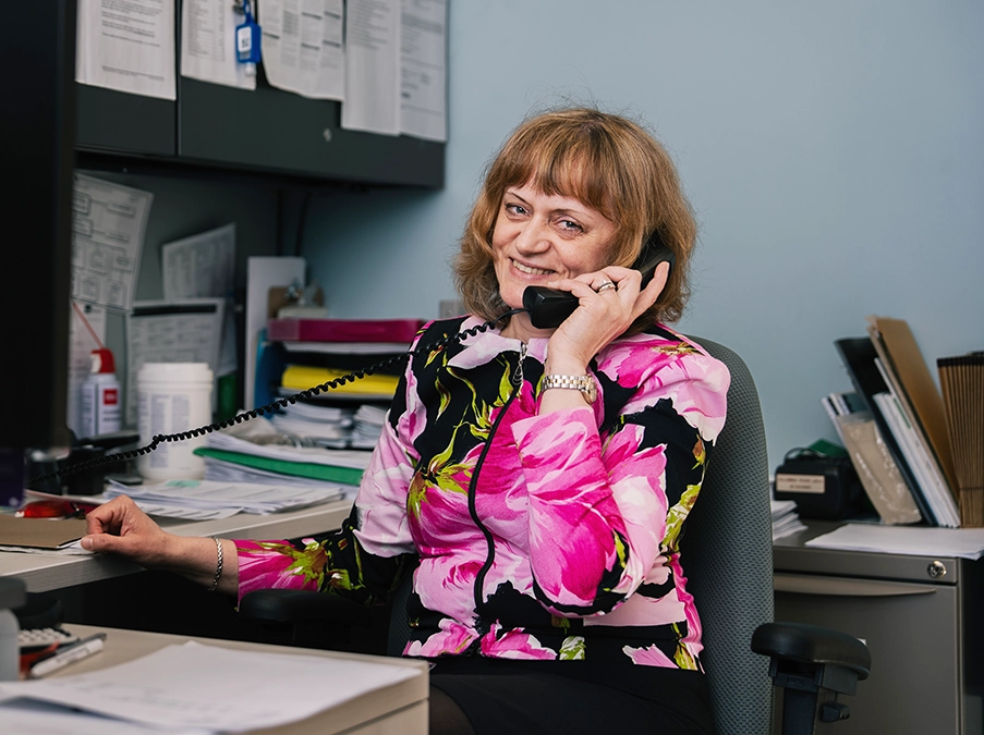 Office administrator seated at a desk, smiling while speaking on a corded telephone, with paperwork and office supplies nearby.