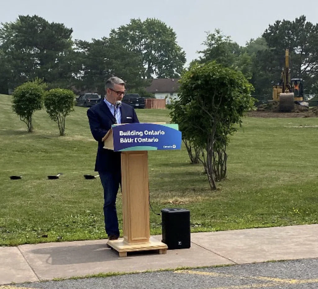 Person standing at a podium with a “Building Ontario” sign during an outdoor announcement, with construction equipment in the background.