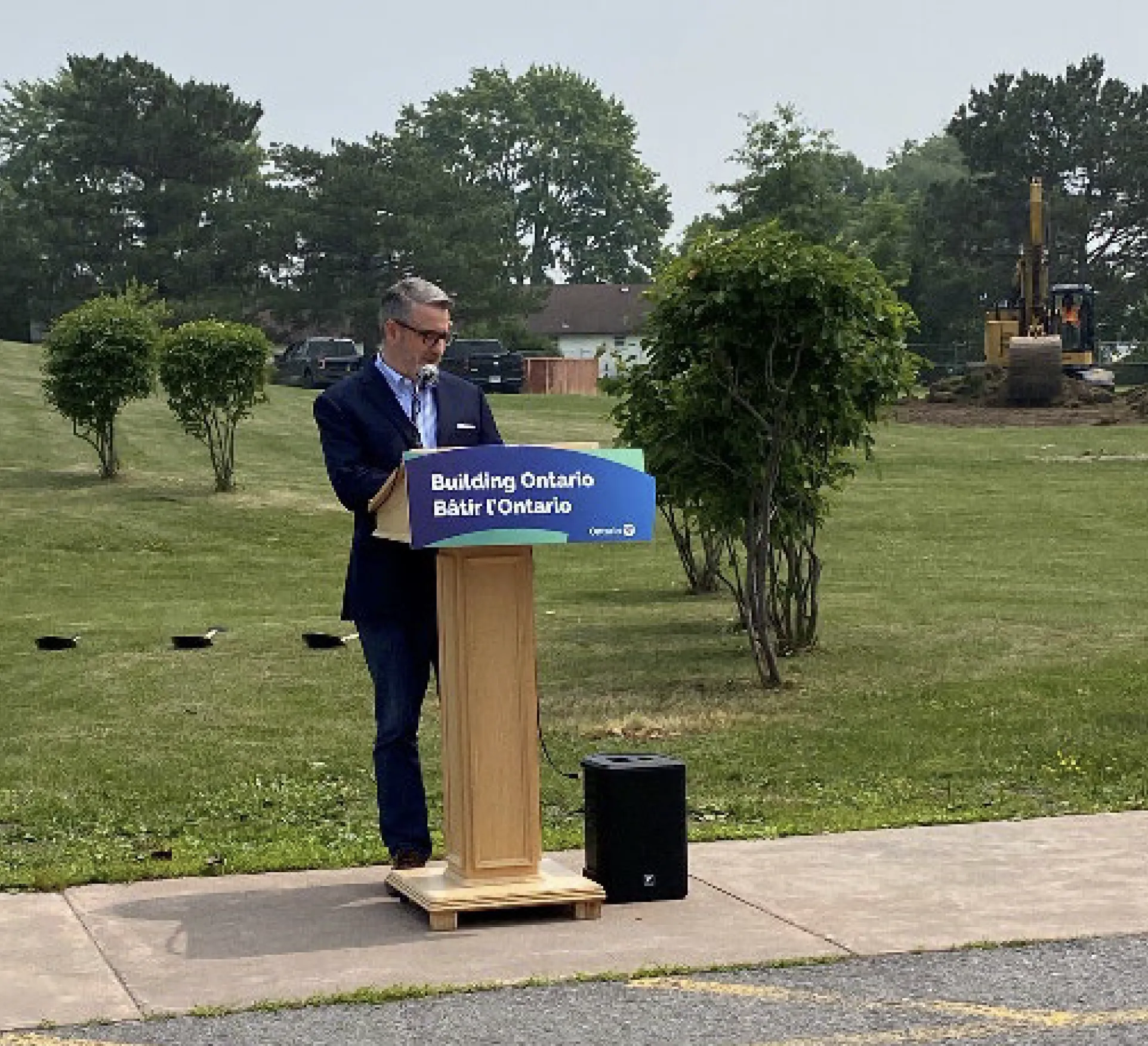 Person standing at a podium with a “Building Ontario” sign during an outdoor announcement, with construction equipment in the background.