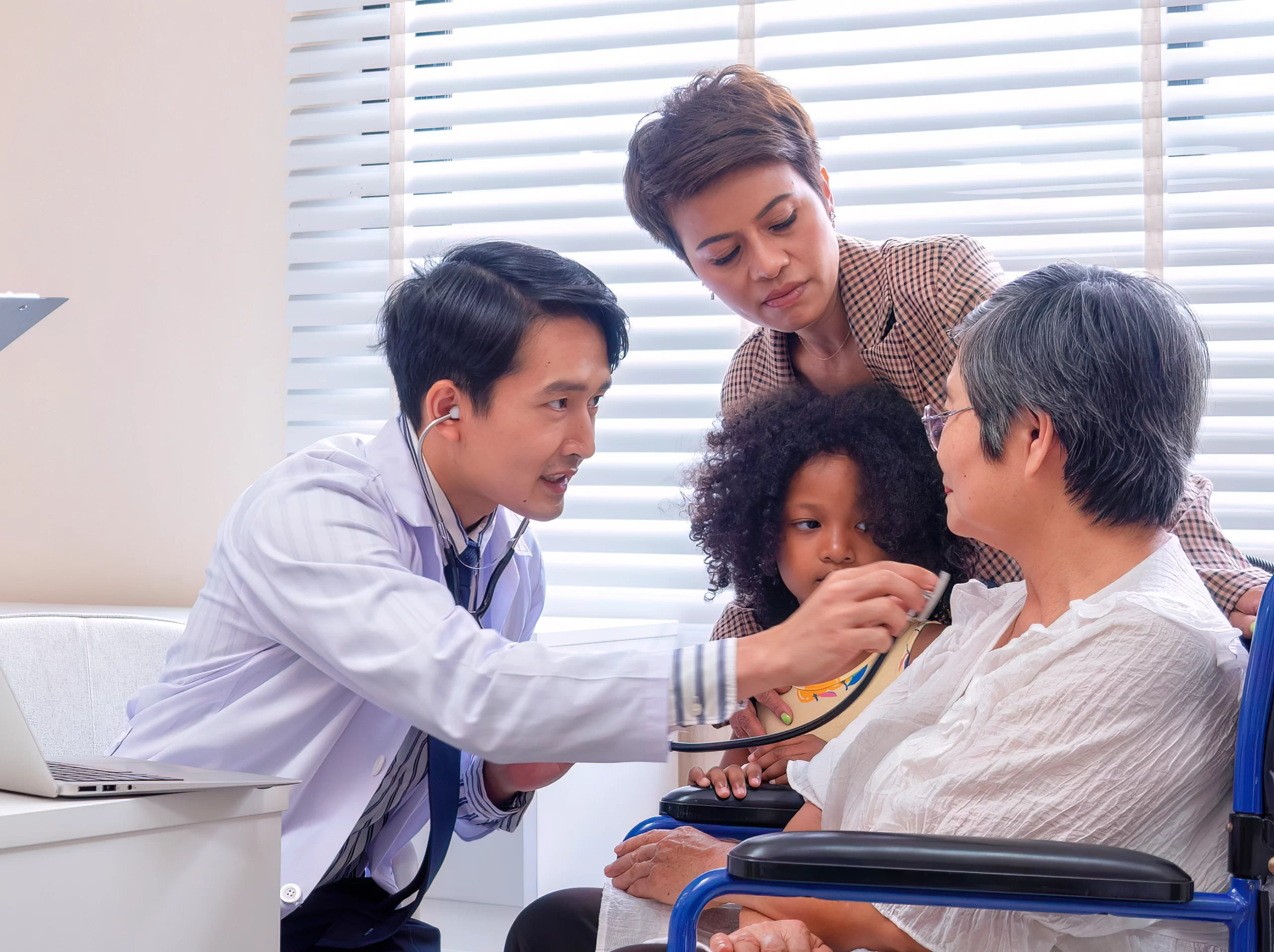 Doctor uses a stethoscope to examine an older patient seated in a wheelchair while a caregiver and child observe.