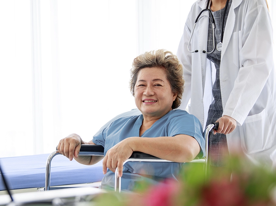 Older patient smiling while seated in a wheelchair as a clinician stands behind them in a hospital room.