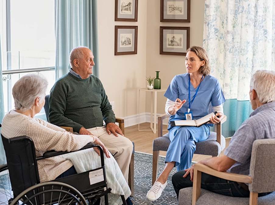 Nurse leading a seated discussion with four older adults in a living room–style care setting.