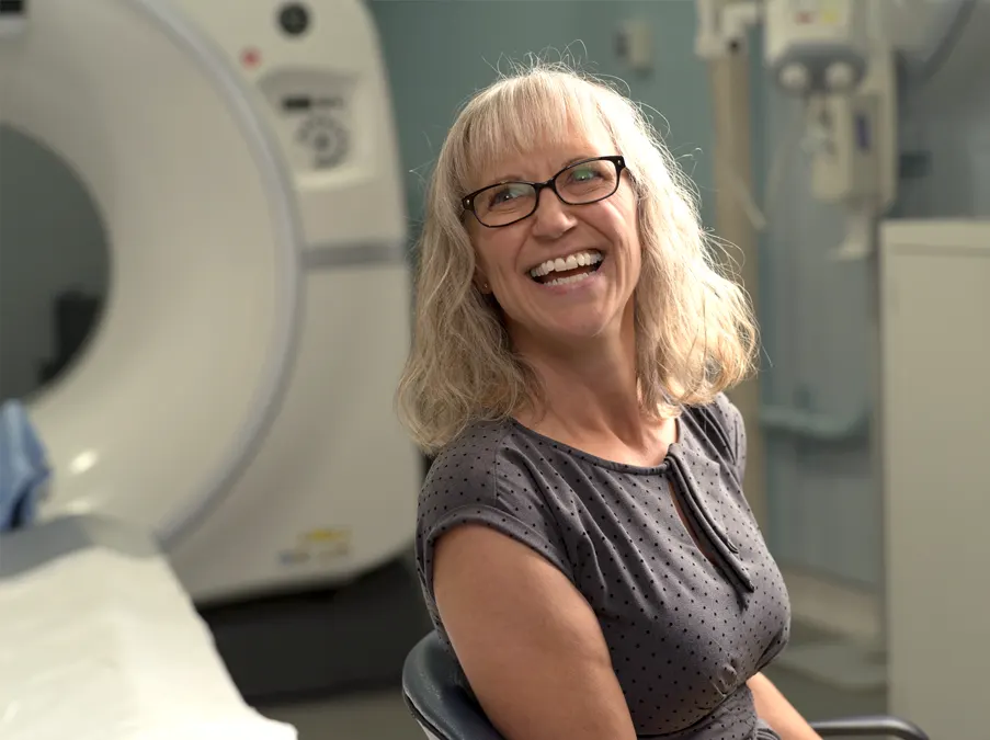Person smiling while seated beside a CT scanner in a hospital imaging room.